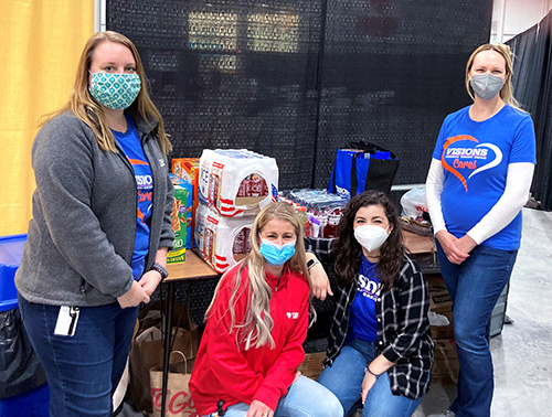 Employees Lindsay, Celeste, Gabriella, and Alexa smile in front of a table with snacks and drinks that Visions donated to workers at the SUNY Broome Vaccination Site.