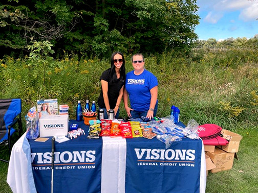 At the 5th hole of the 21st Annual Donald Brubaker Memorial Golf Tournament in Watkins Glen, NY, Visions employees smile at their sponsor table filled with snacks and giveaways for the golfers.