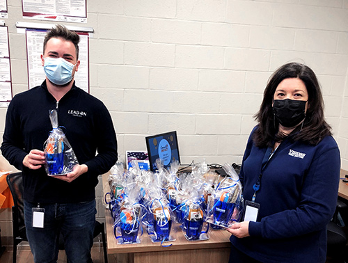 Visions employees, Richie and Donna, smile and hold goodie bags filled with a mug, color changing spoons, Lindt truffles, peppermint bark, biscotti, and assorted hot chocolate to show appreciation for the educators at Leonia High School.