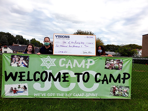 Visions presents the Jewish Community Center of Lehigh Valley with a giant check for $2,500 in front of their “Welcome to Camp” sign.