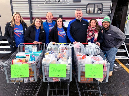 Visions FCU New York Contact Center staff posing with three grocery carts full of food donations for CHOW. There are signs on the carts that read, “Visions FCU Challenges Horizons FCU”