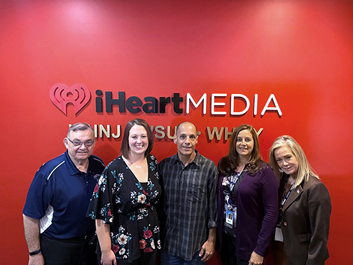 Visions employees stand with three Teachers of the Month in front of a red well and iHeart Media sign.