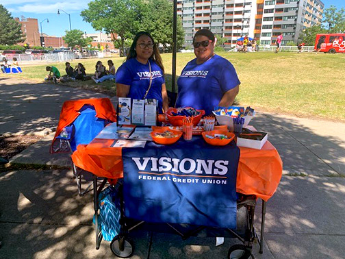Rochester Visions employees stand and smile in the sun at a swag-filled Visions table at Roc Jam Live 2022.