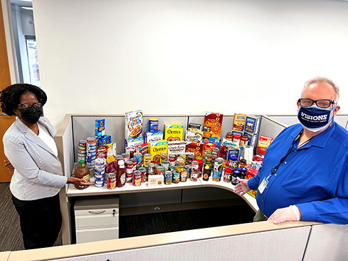 Deiredra (Branch Manager - Visions Saddle Brook Office) and Harry (Member Service Representative - Visions Saddle Brook Office) in NJ show off a table full of food items to be donated to Center for Food Action.