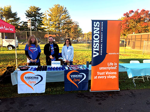 Visions employees pose with their brightly decorated sponsor table at the Thunderbird 5K in Mahwah, NJ. The table is filled with branded goodies that were handed out to all the participants.