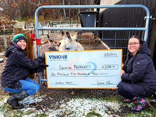 Shannon, Co-founder and Executive Director of Saoirse Pastures, and Megan (from Visions) pictured outdoors on a snowy day, crouching holding a large check in front of two donkeys.