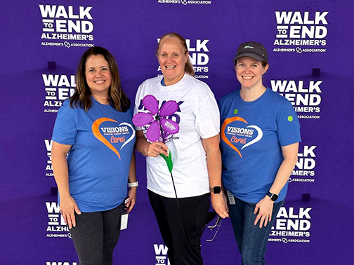 Visions employees Christina, Kelly, and Margaret pictured in front of a purple backdrop with the text: Walk to End Alzheimer’s – Alzheimer’s Association. 
