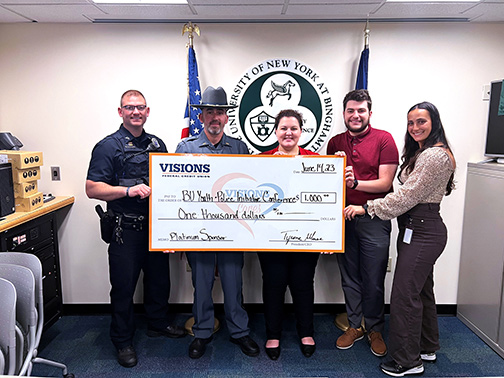 Representatives from the Johnson City Police Department, Binghamton University Police Department, and Visions employees pictured with a large check in front of the seal of the State University of New York at Binghamton.