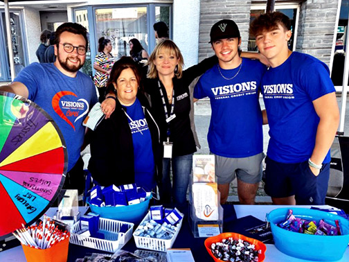 Visions employees posing at a table with various Visions-branded giveaways and a spinning prize wheel. 