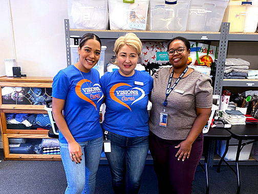 Visions employees Tatiana, Flora, and Desiline pictured in front of a small shelf with clear plastic containers filled to the brim with goods.