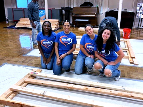 Visions employees Sasha and Tatiana, as well as two Visions high school interns, kneeling and smiling next to a wooden wall frame that they built.