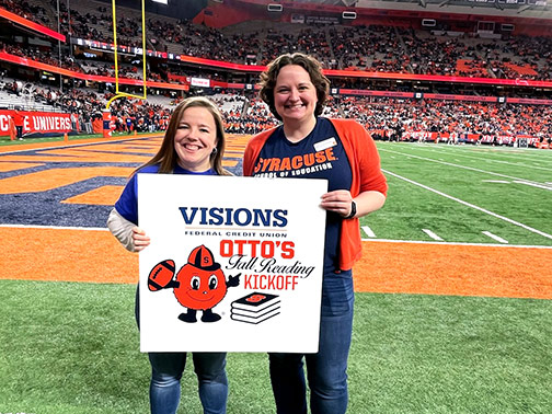 Visions employee, Sarah, and Syracuse University School of Education representative, Allison, standing on the Syracuse football field, smiling, and posing with a white sign that says Visions Federal Credit Union: Otto’s Fall Reading Kick Off. 