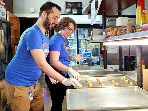 Visions employees Raul and Clairissa happily rolling balls of cookie dough onto a baking sheet in a kitchen.