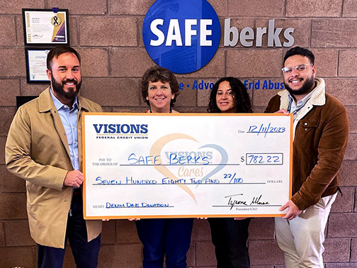 Visions employees Derek, Millie, and Gustavo pictured with Safe Berks Director of Development, Mindy McIntosh. They are pictured in front of a brick wall with various awards and the Safe Berks logo, all while smiling and holding a large check.