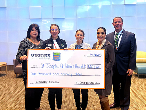 Visions employees Lori and Tatiana, as well as representatives from St. Joseph’s Children Hospital, smile and pose with a large check in an open lobby area.
