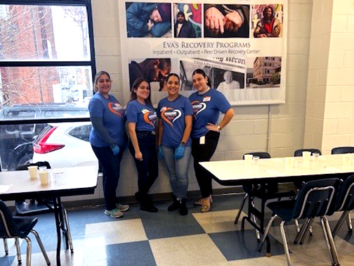 Visions employees Julia, Carilyn, Jurgena, and Tatiana standing and smiling in front of an Eva’s Village banner in a cafeteria.