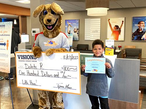 Dakota stands next to Kirby the Kangaroo holding a Little Hero Certificate. Kirby is holding a giant $100 check.